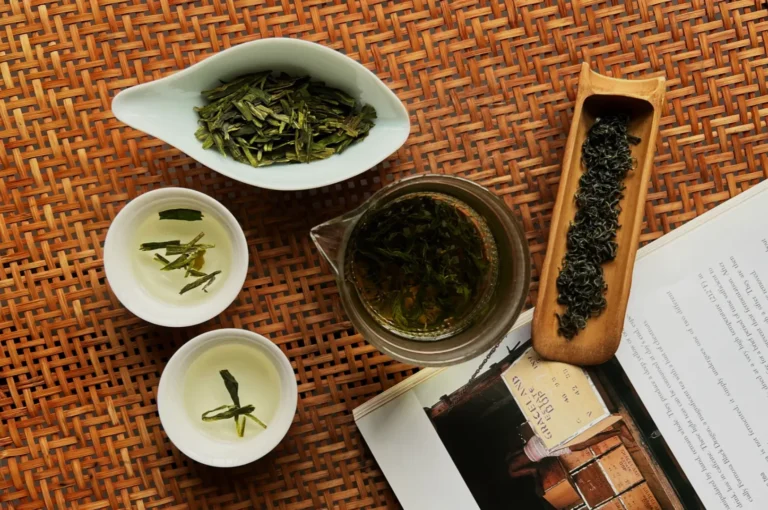 A flat lay of tea leaves, a teapot with brewed tea, two small cups of tea, and a magazine on a woven mat.