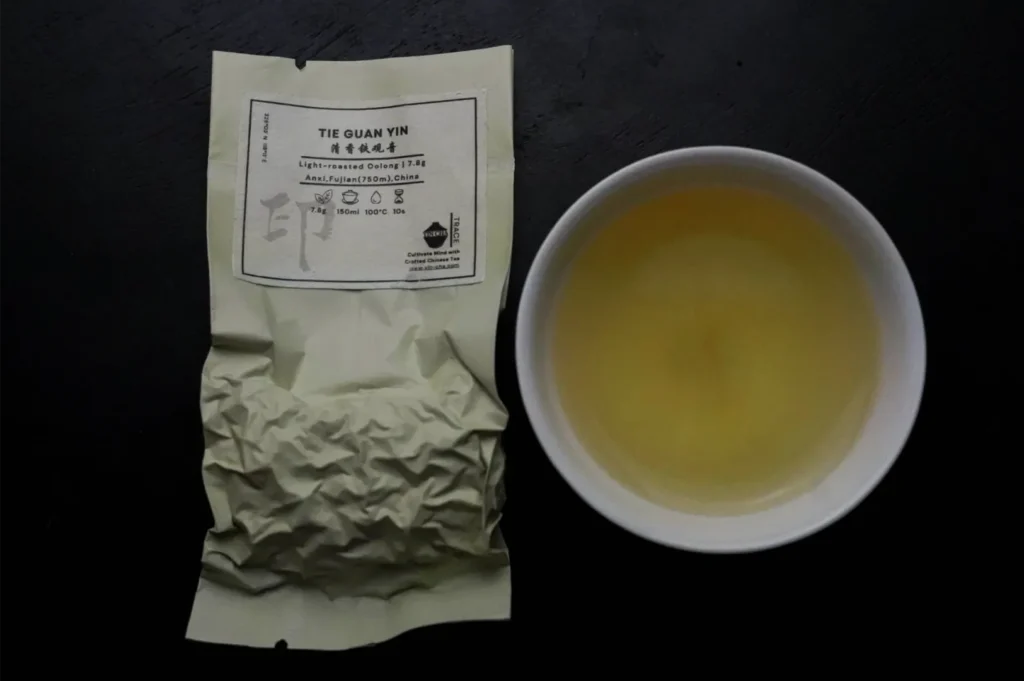 A crinkled beige tea pouch labeled “Tie Guan Yin” sits beside a white bowl of golden tea on a dark surface, creating a calm, minimalist presentation.