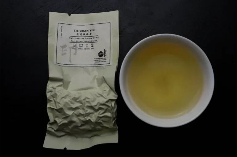 A crinkled beige tea pouch labeled “Tie Guan Yin” sits beside a white bowl of golden tea on a dark surface, creating a calm, minimalist presentation.