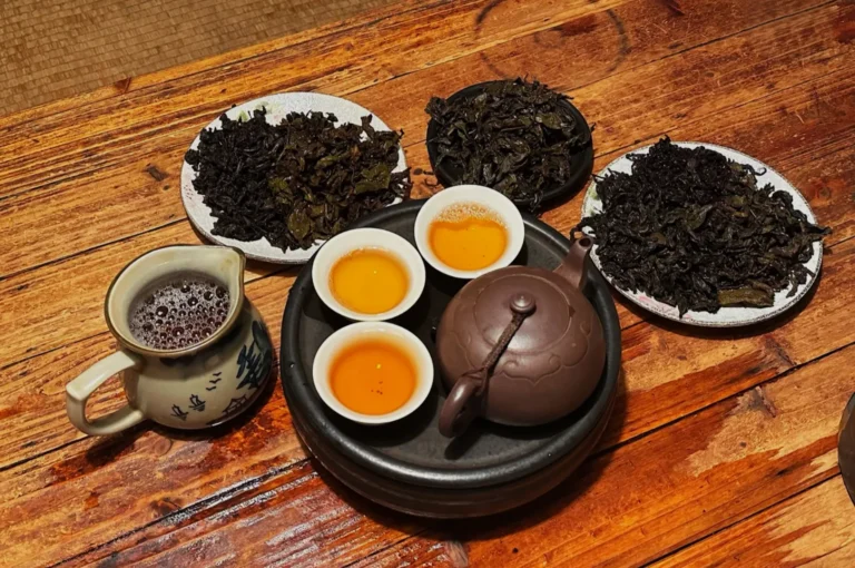 A teapot with three tea cups filled with tea, surrounded by plates of loose tea leaves on a wooden table.