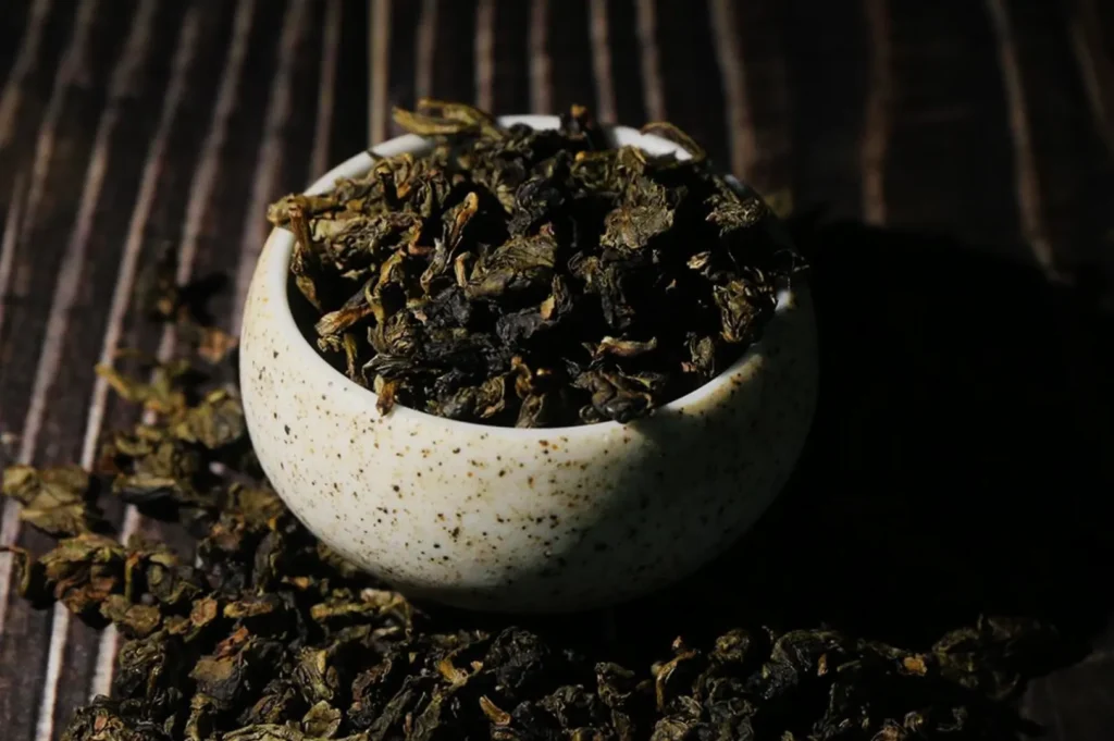 A speckled ceramic bowl brimming with loose dried tea leaves, surrounded by scattered leaves on dark wood, creating a rustic, moody atmosphere.