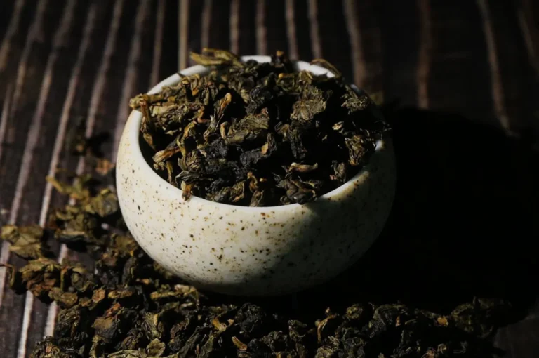 A speckled ceramic bowl brimming with loose dried tea leaves, surrounded by scattered leaves on dark wood, creating a rustic, moody atmosphere.
