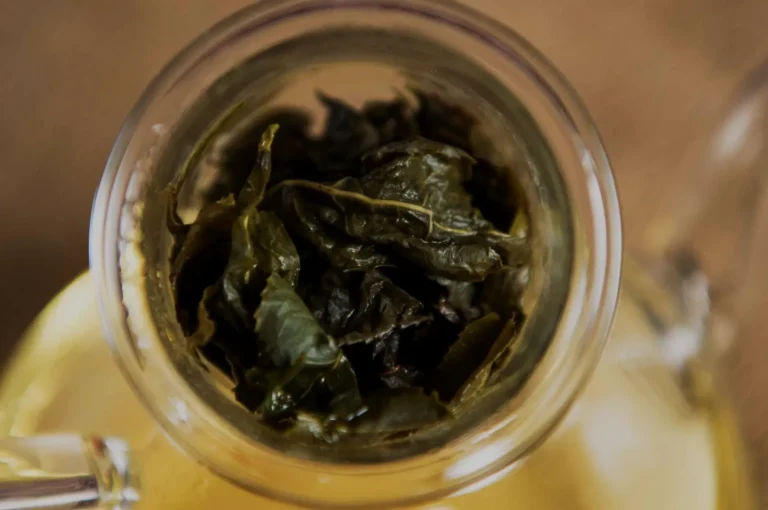 Top-down view of a glass jar filled with dark, crinkled green leaves, surrounded by warm amber liquid and soft light, creating a rustic mood.