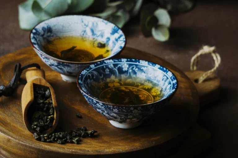 Two ornate bowls filled with tea, placed on a wooden tray alongside a wooden scoop holding loose tea leaves. Eucalyptus leaves are softly blurred in the background.