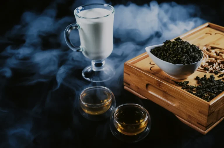 A glass of milk, two small cups of tea, and a bowl of dried tea leaves on a wooden tea tray with smoke in the background.