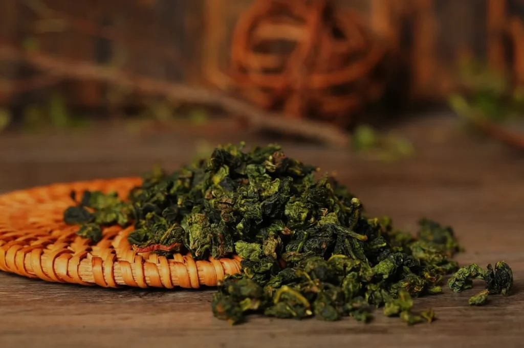A pile of dried green tea leaves on a woven mat with a blurred background of twigs and leaves.