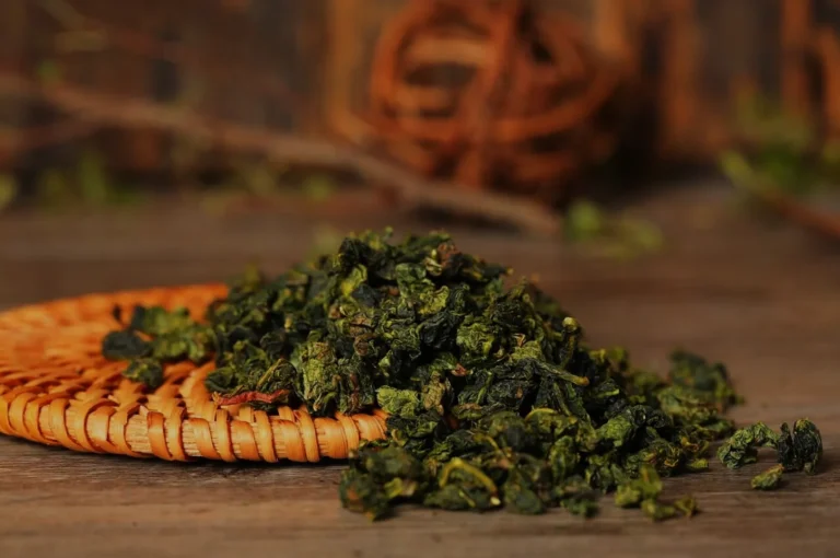 A pile of dried green tea leaves on a woven mat with a blurred background of twigs and leaves.