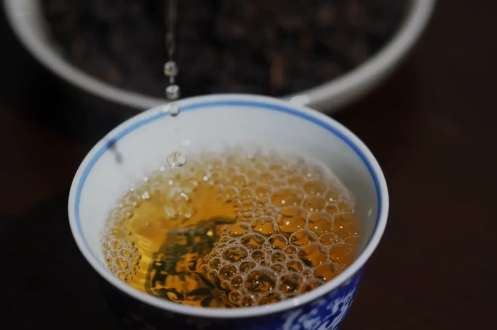 A close-up of a cup filled with bubbling yellow tea, with a blurred background of dark tea leaves.