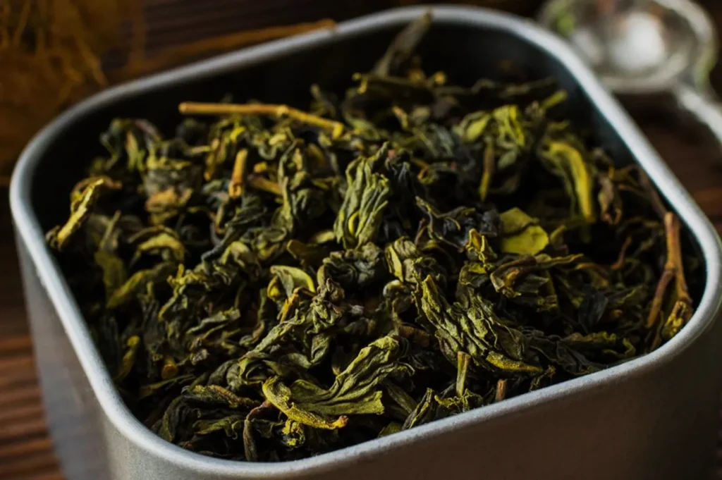 A close-up of dried green tea leaves in a metal container.
