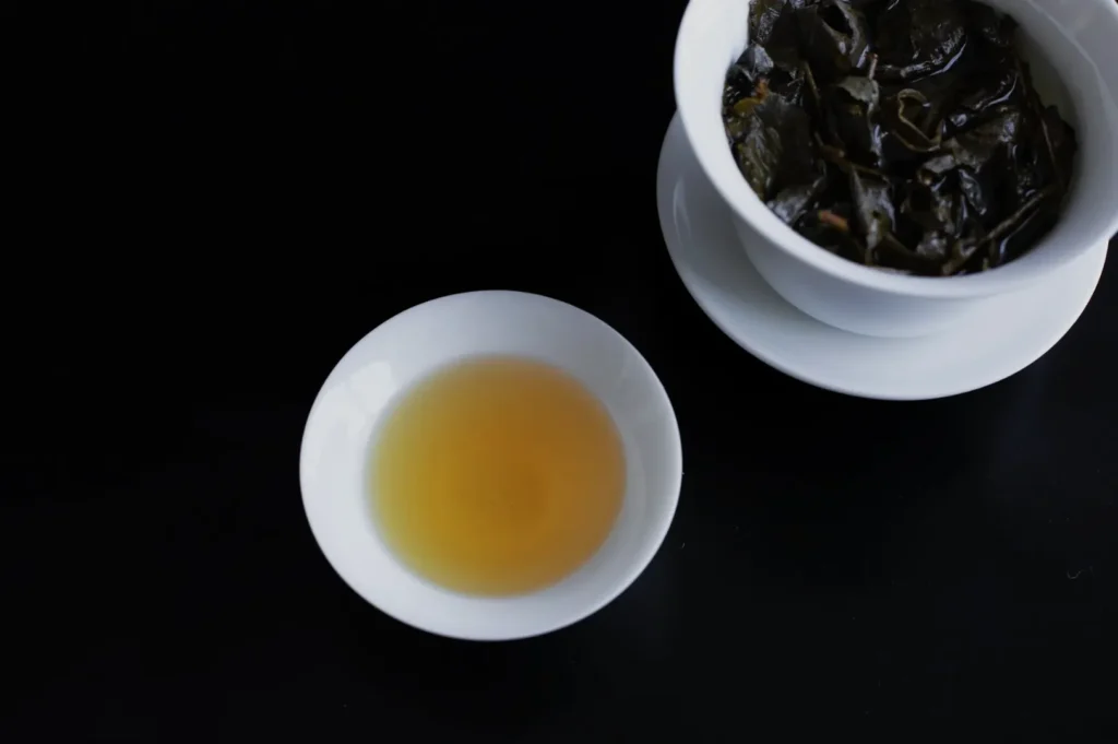A cup of brewed tea next to a bowl of dark tea leaves on a black surface.