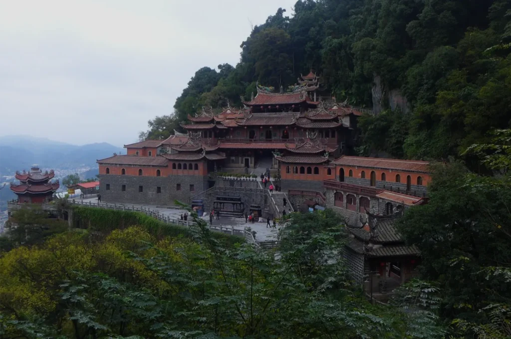 A tiered Chinese temple complex clings to a misty mountainside, with ornate red roofs, stone walls, visitors, and lush green forest.