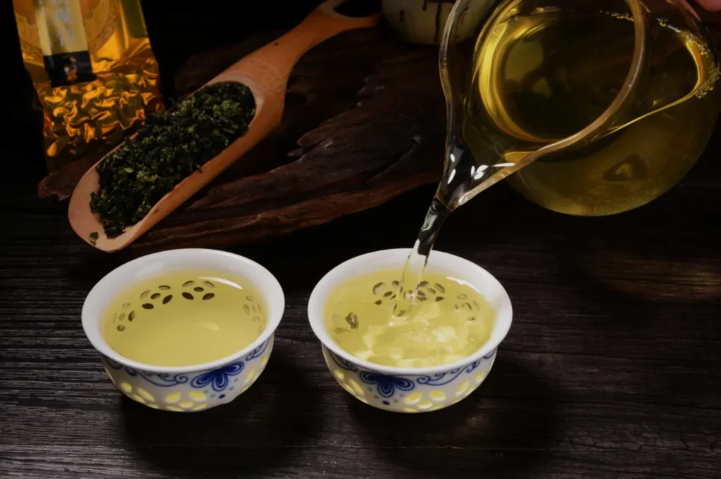 Golden tea pours from a glass pitcher into a blue-and-white porcelain cup; a second cup and wooden scoop of loose tea sit on a dark table, evoking calm.