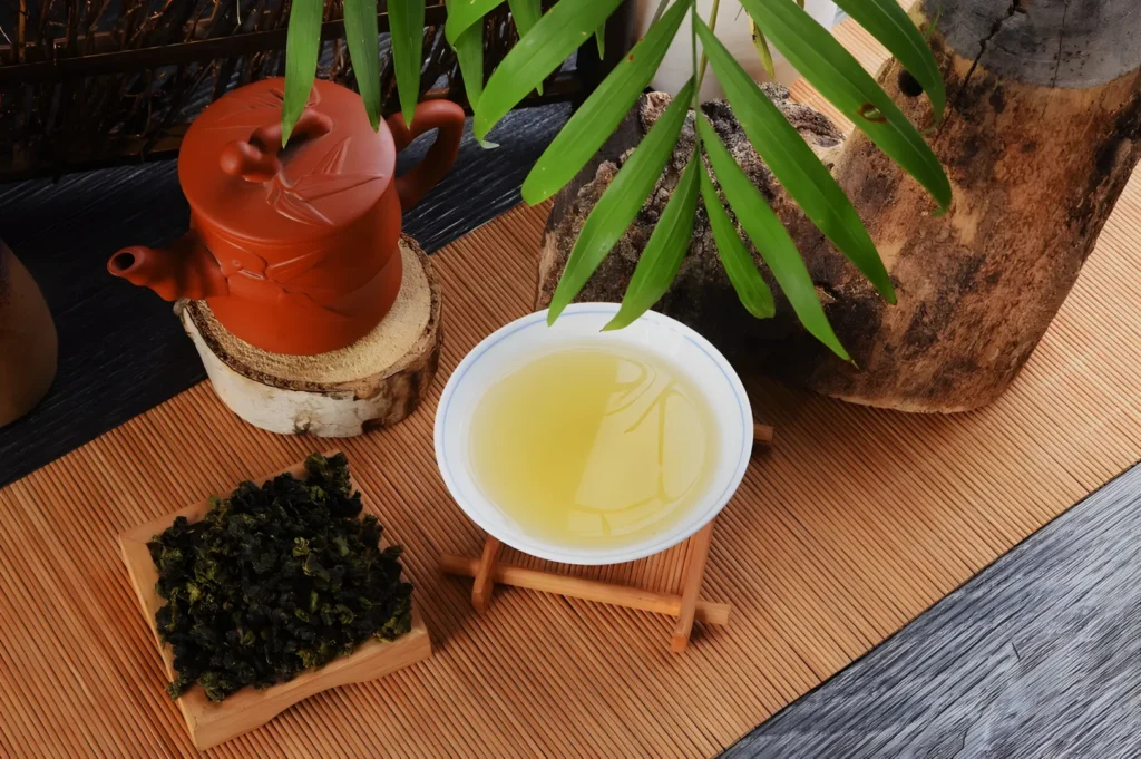 A tea setup featuring a clay teapot, a bowl of light yellow tea, and a small dish of loose tea leaves, surrounded by natural elements like a wooden surface and green leaves.