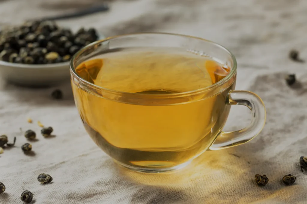 A clear glass cup filled with golden-yellow tea, placed on a light fabric surface with scattered tea leaves and a small bowl of loose tea in the background.