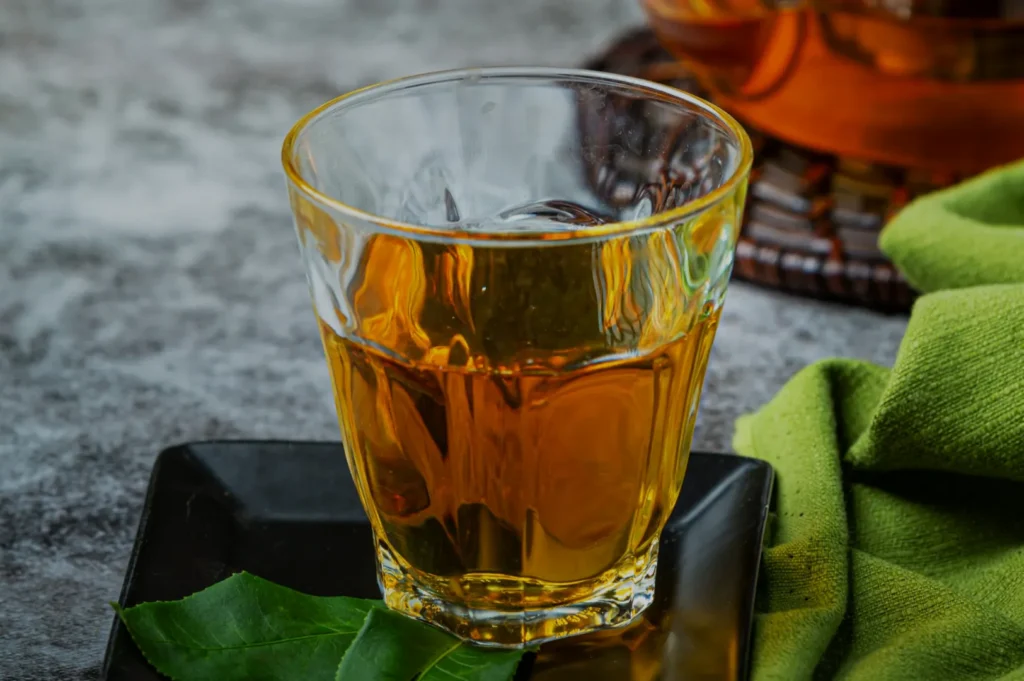 A glass of amber-colored tea sitting on a black plate, with a green cloth and tea leaves in the background.