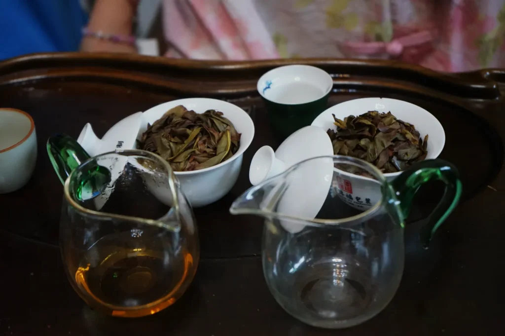 A wooden tea tray with multiple small bowls of loose tea leaves and glass teapots, one containing brewed tea.