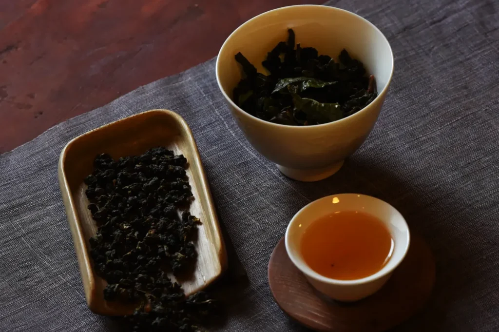 A bowl of loose tea leaves next to a small cup of brewed tea, all set on a textured cloth.
