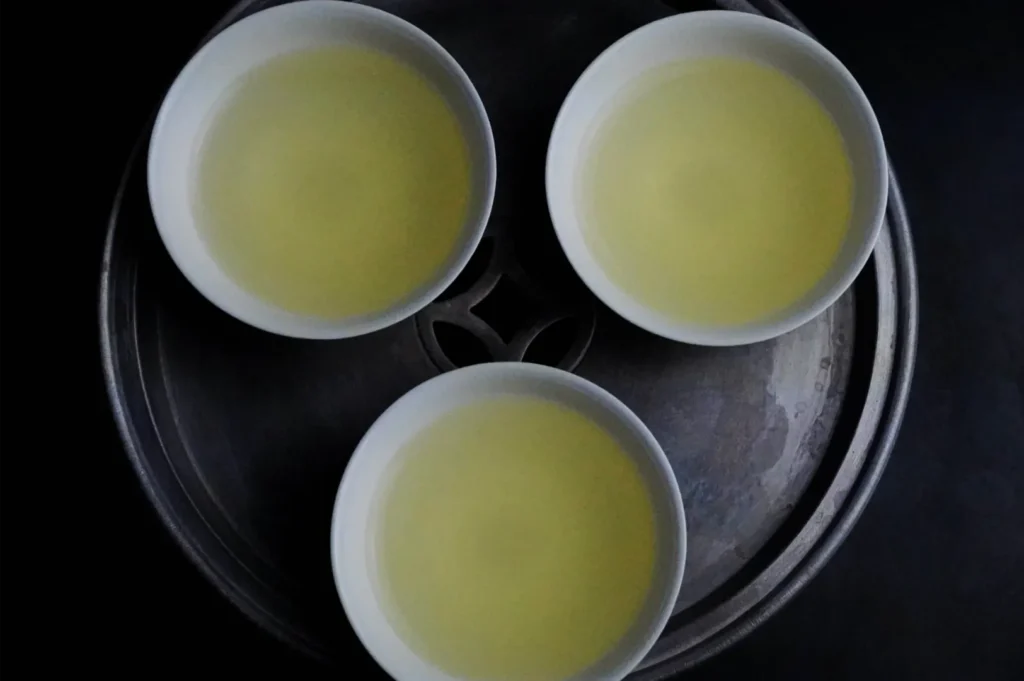 Overhead view of three white bowls filled with pale yellow liquid arranged in a triangle inside a dark metal pot, creating a quiet, minimalist mood.