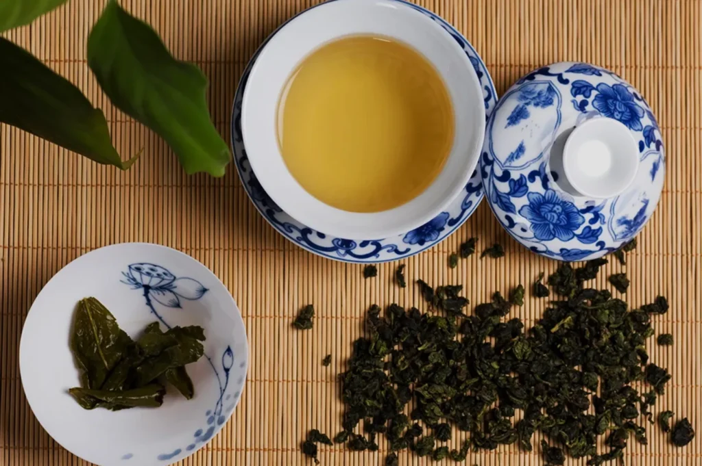 A flat lay of a tea setup, featuring a cup of golden tea, a small dish with green tea leaves, and loose dried tea leaves scattered on a bamboo mat, accompanied by a small white lid.