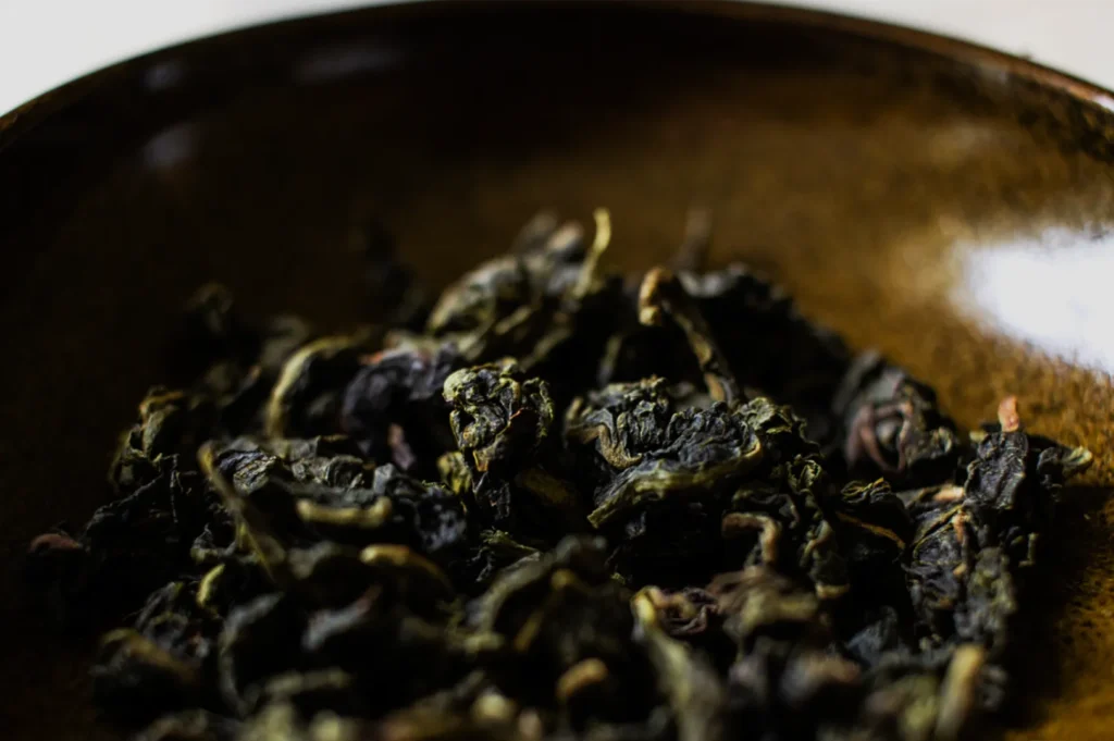 Close-up of curled dark green loose-leaf tea in a glossy brown ceramic bowl, creating a rustic, earthy, moody impression.