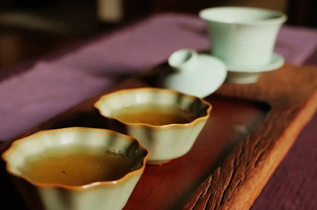 Three cream-colored ceramic tea cups with scalloped, amber-edged rims sit on a wooden tray beside a white teapot, creating a warm, intimate tea-time scene.
