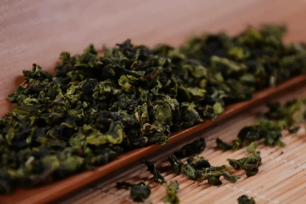Close-up of curled green tea leaves piled on a wooden scoop, with scattered leaves on a warm bamboo surface, creating a rustic, earthy mood.
