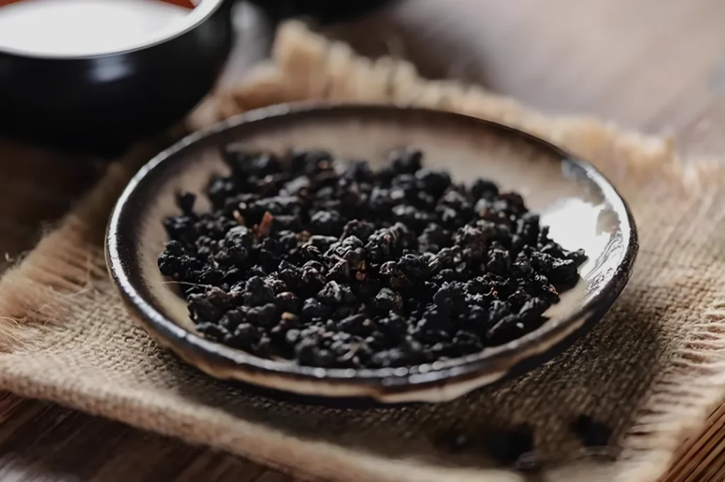 A plate filled with dark, dried fruits or herbs, placed on a textured fabric surface.