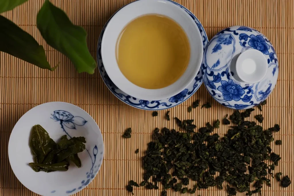 A traditional tea setup with a blue and white bowl containing brewed tea, a matching lid, and a small dish of tea leaves, arranged on a bamboo mat.