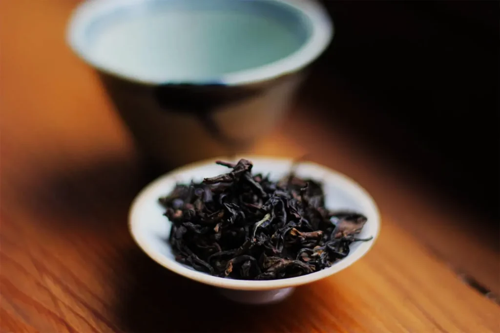 A small dish of dark, dried tea leaves next to a tea cup on a wooden surface.
