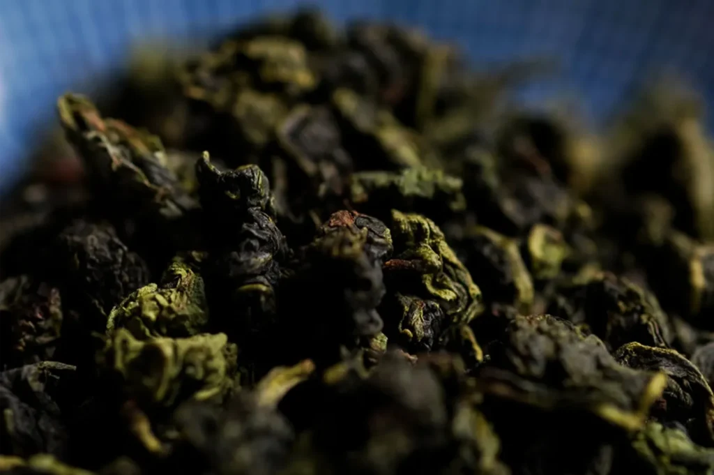 Close-up of curled, dried tea leaves in mottled green and brown, piled in shallow focus against a blurred blue background; the moody lighting feels earthy and rustic.