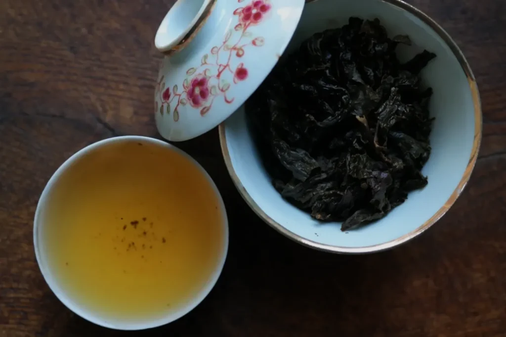 A bowl of tea leaves next to a cup of brewed tea, all placed on a wooden surface.
