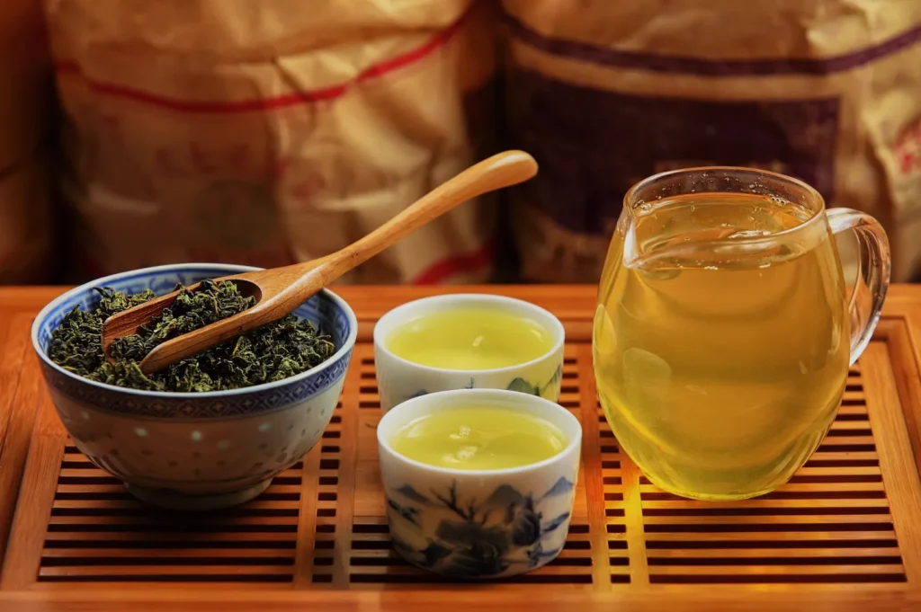 Warm still life of loose-leaf tea in a patterned bowl with wooden scoop, two cups of pale tea, and a glass pitcher on a bamboo tray.