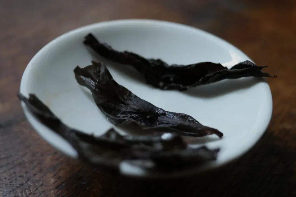 Several wrinkled, dried black chili peppers rest on a white ceramic plate atop a dark wooden table, creating a rustic, moody still life.