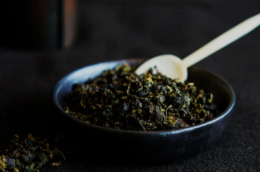 A small black bowl filled with loose dried tea leaves, accompanied by a white spoon resting on the surface.
