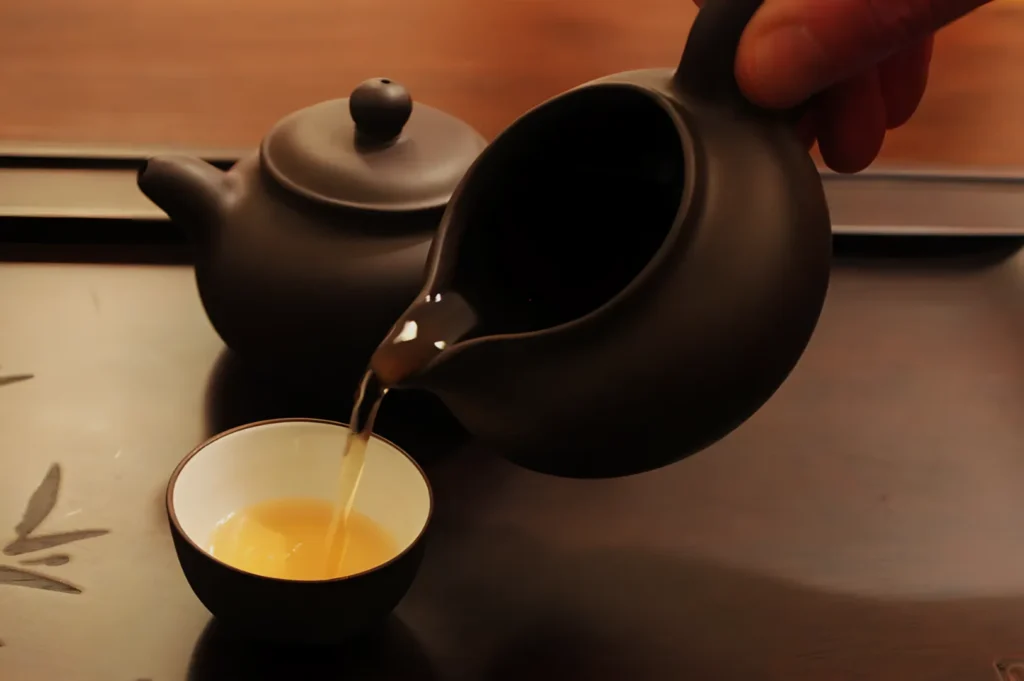 A person pouring tea from a dark teapot into a small cup on a wooden surface.