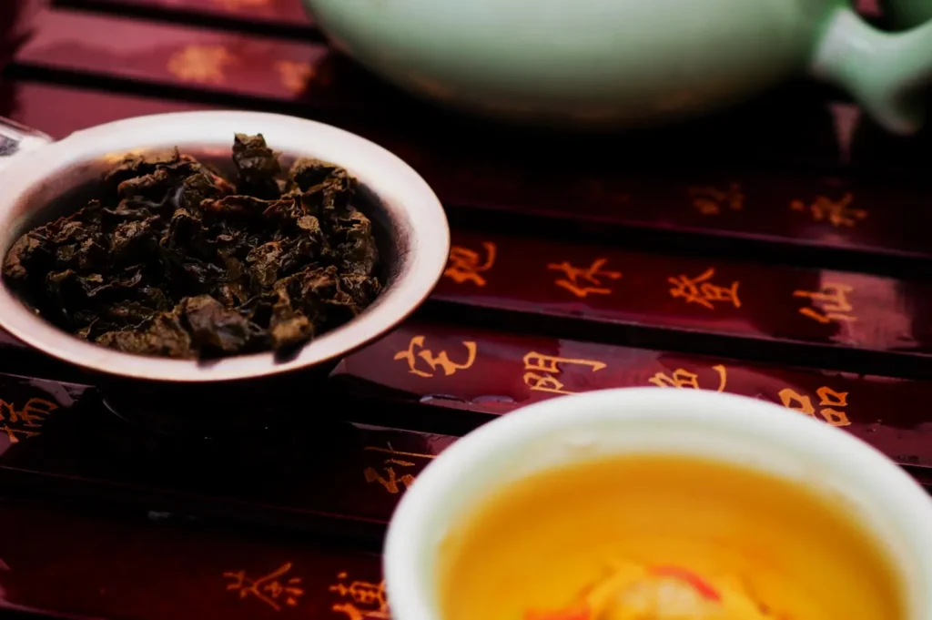 A close-up of a bowl of dry tea leaves next to a cup of brewed tea on a decorative wooden surface with Chinese characters.