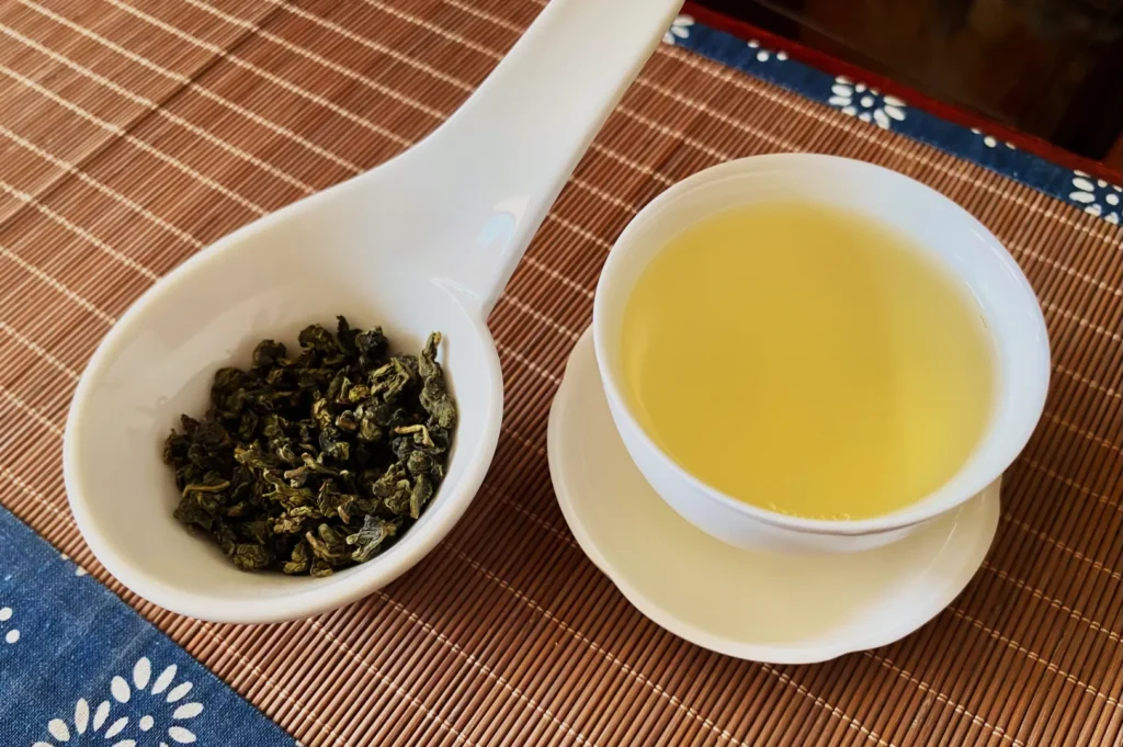 A bowl of brewed green tea next to dry green tea leaves on a bamboo mat.