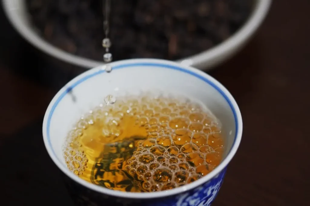 A cup of tea with bubbles, set against a blurred background of dark tea leaves.