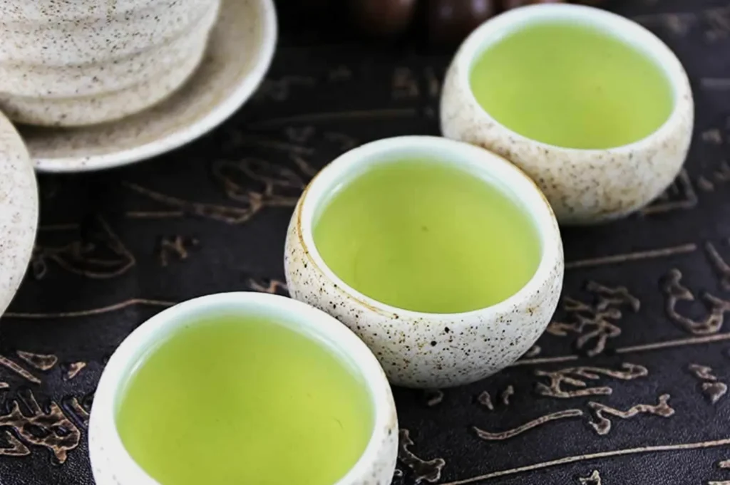 Three speckled white ceramic cups filled with pale green tea sit on a dark patterned tray beside a matching teapot, creating a calm, inviting mood.