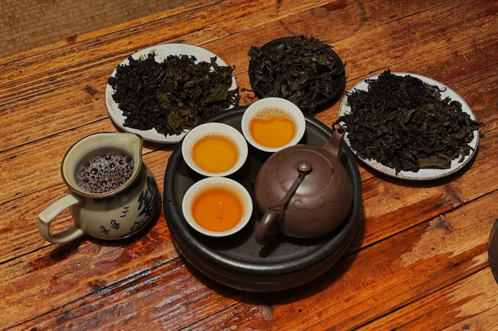 A wooden table with a teapot and small cups of tea. There are plates of loose tea leaves around the teapot.