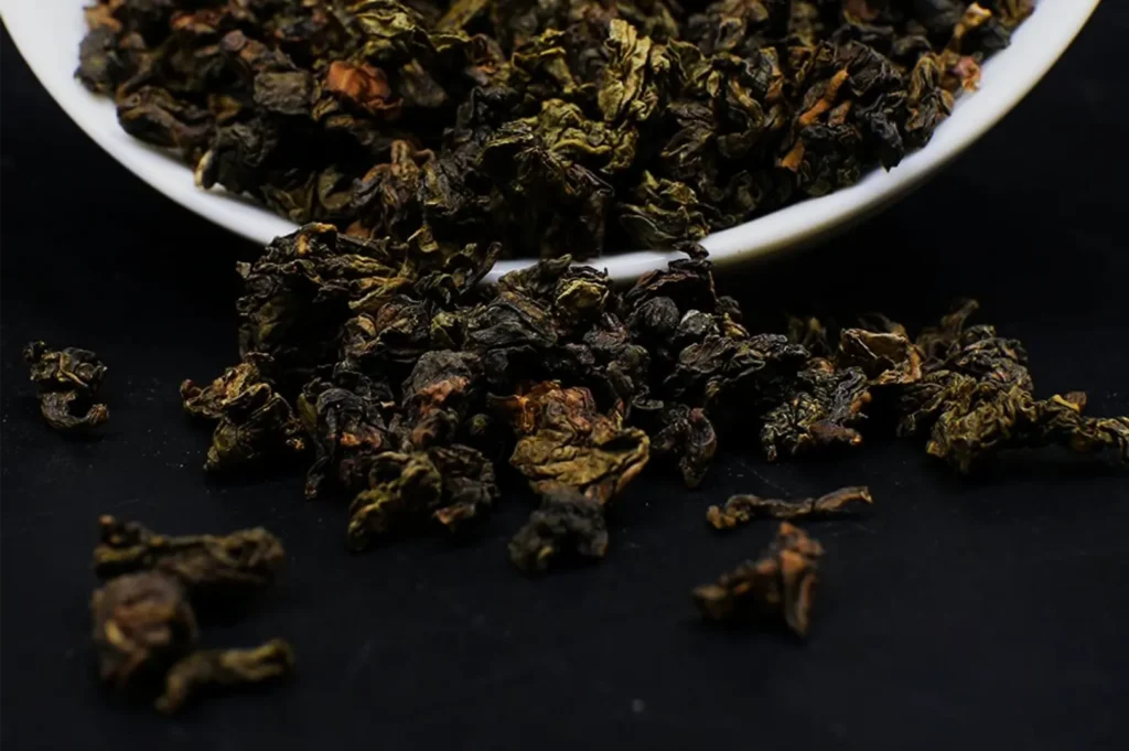 Close-up of loose-leaf dark tea spilling from a white bowl onto a black surface, with curled brown-green leaves and an earthy, rustic tone.