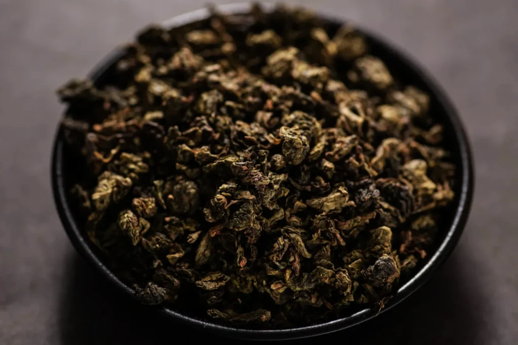 Black bowl filled with dense, crinkled dried herbs or tea leaves, photographed from above against a dark gray background, creating a rustic mood.