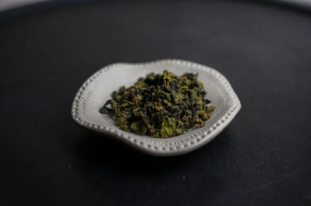 A small white dish containing loose green tea leaves on a dark surface.
