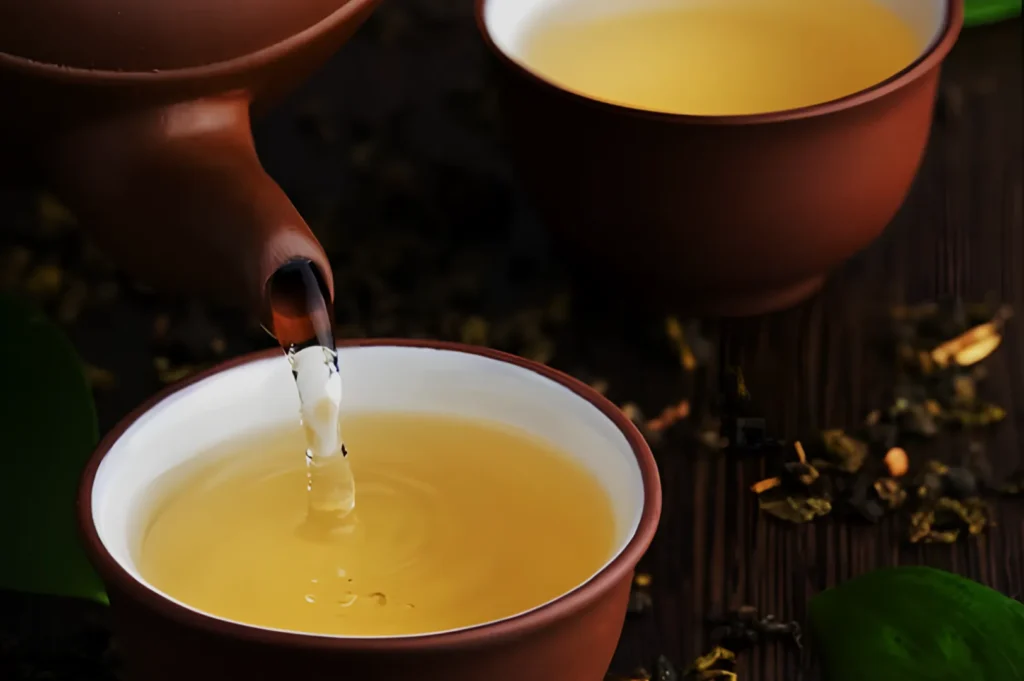 A teapot pouring yellow tea into a small brown cup, surrounded by loose tea leaves and green leaves on a wooden surface.