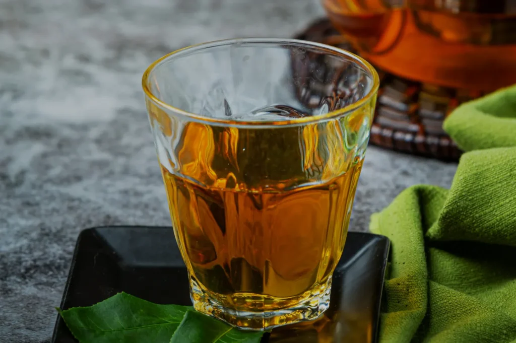 A glass of amber-colored tea on a black plate, with a green cloth and some leaves in the foreground.