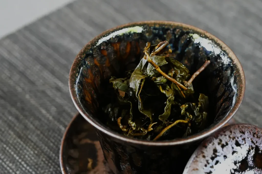 A dark, textured cup filled with dried green tea leaves, sitting on a saucer.