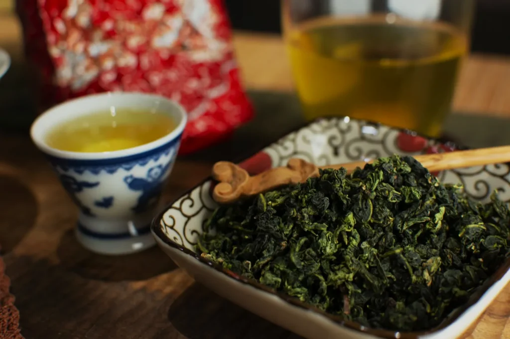 A bowl of loose green tea leaves alongside a small cup of tea, with a glass teapot in the background.