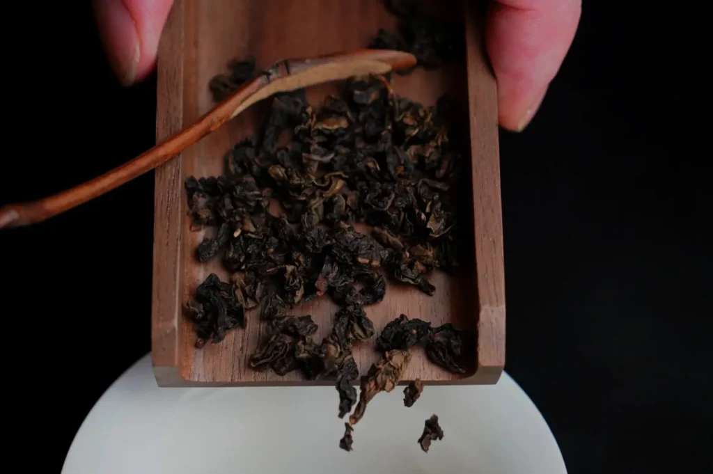 A wooden scoop holding dried tea leaves above a round white container.