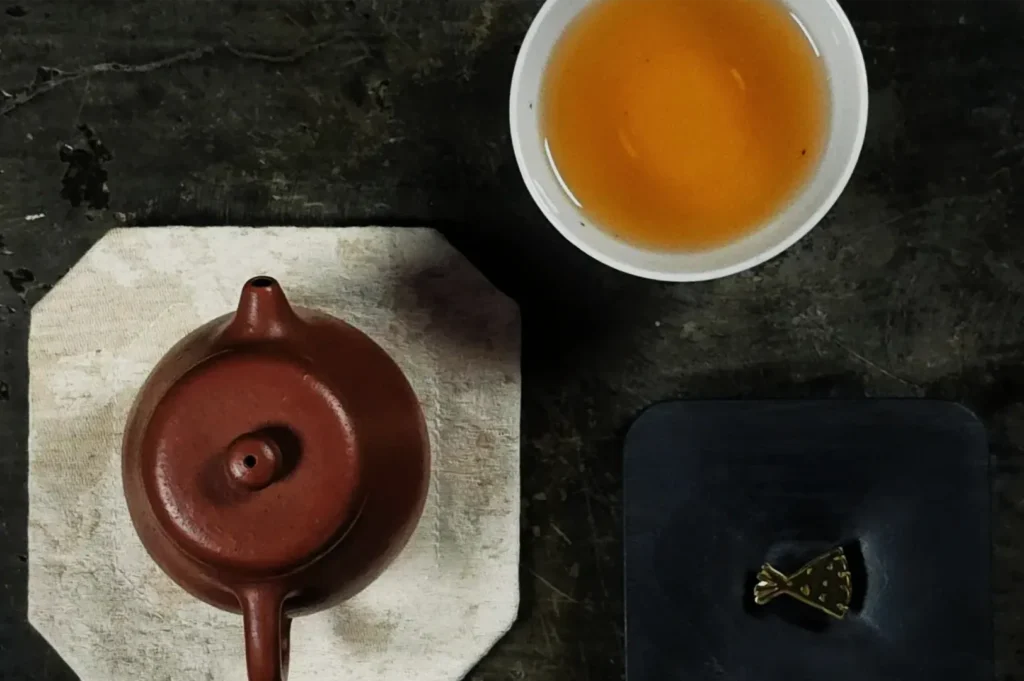 Overhead view of a reddish clay teapot on a pale stone board, beside a cup of amber tea and a small gold-toned tea tool; calm tea ritual.