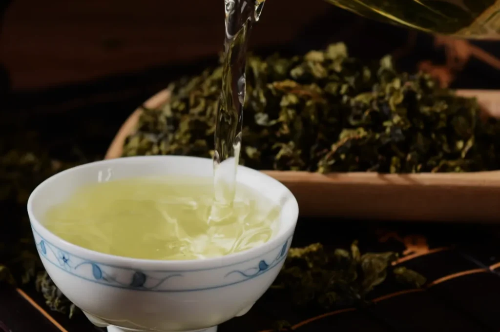 A cup of green tea being poured from a teapot, with loose tea leaves in the background.